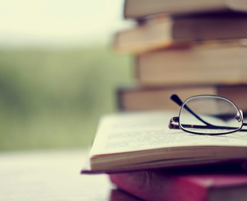 stack of vintage books on the table, glasses and an open book. in the background park forest